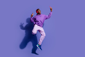 Obraz premium Joyful young man in purple hoodie jumping and listening to music, using smartphone and headphones, on violet background