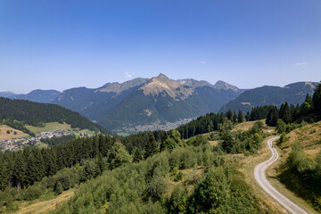 Aerial mountainous landscape with meandering path through lush green French Alps valley tourist destination winter sports near Les Gets village with rocky mountain top against a clear blue sky. 
