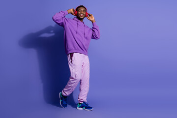 Young stylish man in trendy purple outfit enjoying music on headphones with a cheerful smile against a vibrant backdrop