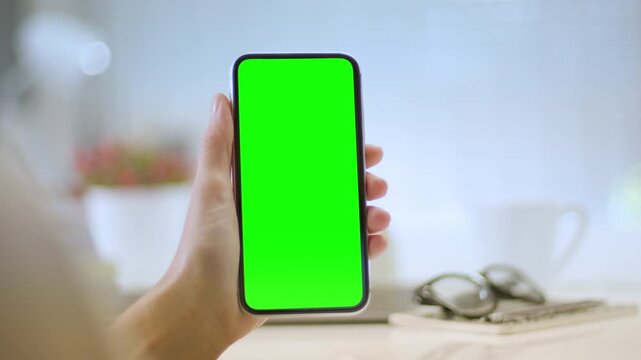 Close Up Of Hand Holding Smartphone With Green Screen In Bright Modern Office, Representing Mobile Technology, App Design, Mockup, User Interface And Digital Innovation.
