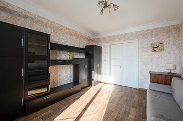 bright living room with dark furniture set against floral wallpaper. Sunlight streams across the wooden floor. The room features a couch, wardrobe, and a decorative chandelier