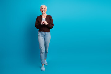 Stylish senior woman with short grey hair holding a smartphone in casual jeans and cardigan against blue background