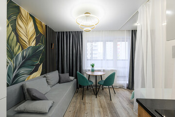 cozy living space with a sofa, round table, two green chairs, and a leaf-patterned accent wall.  Bright lighting and curtains create a modern, inviting feel