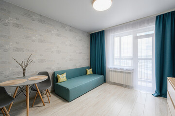 cozy living space features a teal sofa with patterned cushions, a round table with gray chairs, light wallpaper, and a bright window with blue curtains