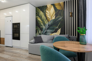 modern living space with a kitchen corner, featuring a grey sofa, round wooden table, turquoise chairs, and a botanical wall mural