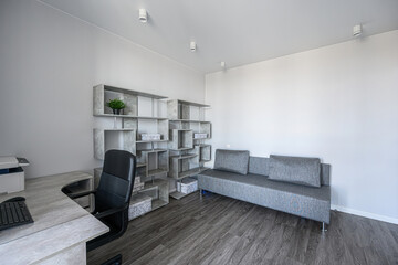 modern, minimalist office space with a gray sofa, desk, bookshelf, and dark wood floor. The room is bathed in neutral tones and natural light