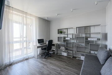 Bright, modern room featuring a desk with computer by a window with sheer curtains.  A gray bookcase and sofa are present, creating a minimalist workspace