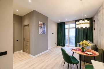 Cozy apartment interior featuring a dining area with an oval table, velvet chairs, and green curtains. The room has modern lighting, light wood floors, and a balcony view