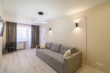 Cozy living room with a grey sofa, wooden floors, and beige walls. Modern lighting and curtains create a comfortable space
