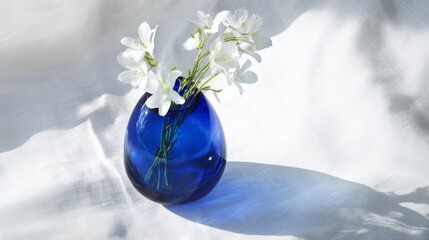 Soft sunlight highlighting cobalt blue glass vase with white blooms, casting gentle shadows on pristine linen surface, embodying tranquil elegance