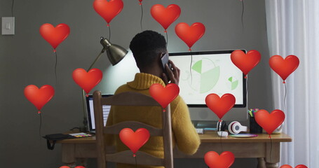 Talking woman wearing mustard yellow sweater holding smartphone in office with red heart balloons