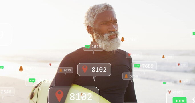 Preparing senior man wearing black wetsuit holding surfboard on beach, with floating social icons - Powered by Adobe