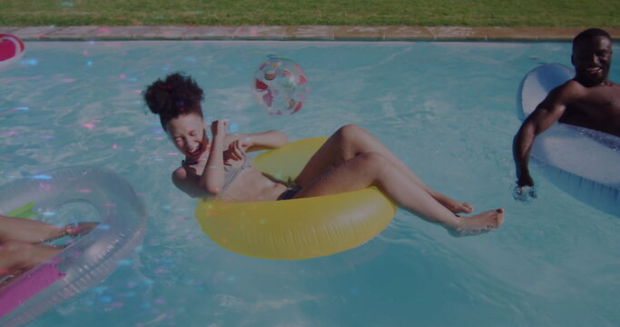 Laughing woman wearing swimsuit in yellow tube at poolside, with white tube, pink tube, beach ball
