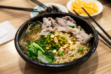 Bowl of hot noodle soup with sliced pork, peanuts, pickled vegetables and fresh greens in savory broth served in a black bowl on wooden table