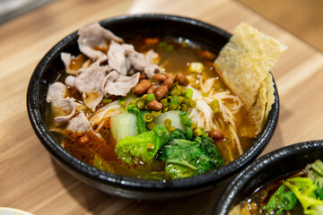 Hot bowl of spicy rice noodles with sliced pork, peanuts, fresh vegetables and crispy tofu skin in chili broth served in black bowl
