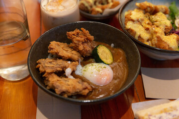 Fried chicken pieces with soft boiled egg and zucchini on rice, served in black bowl with rich brown curry sauce in a cozy restaurant setting