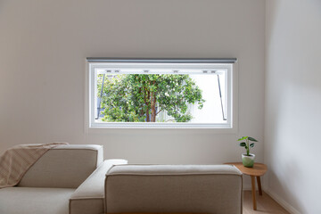 Minimalist living room with neutral tones, white wall, cozy armchairs and side table, open window view of lush green garden tree outside