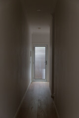 Narrow hallway with wooden floor and light streaming through frosted glass door, creating moody contrast in minimalist home interior design