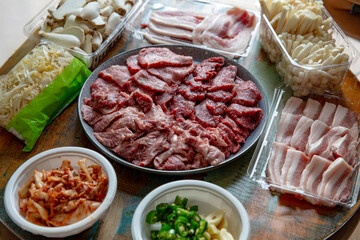 Top down view of fresh Korean BBQ spread featuring sliced beef, pork belly, mushrooms, kimchi, garlic, chilies and bean sprouts on rustic table