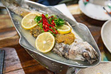 Whole steamed fish with lemon slices, chili and herbs served in metal fish-shaped dish, Thai cuisine presented in rustic restaurant table setting