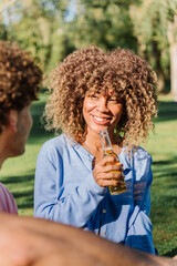 Fototapeta premium Curly-haired woman laughing, gripping beer bottle while enjoying sunny park hangout with close friend, radiating happiness during leisurely outdoor gathering