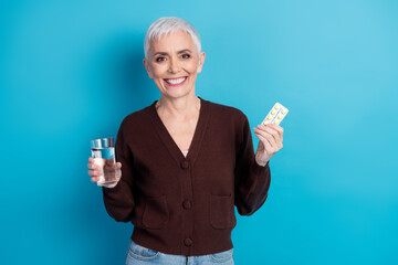 Elderly woman holding a glass of water and a pill pack against a blue background, showcasing healthy lifestyle and self care.