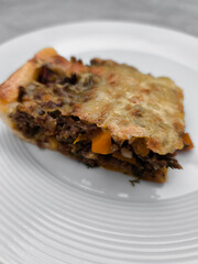 Slice of delicious homemade meat pie with melted cheese and vegetables on white plate