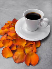 White cup of coffee with orange rose petals on gray table