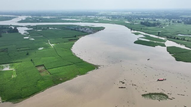 Bogura, Bangladesh - 22 August 2025: Aerial view of a wide river snaking through lush green fields, adjacent to a village with tightly packed houses.
