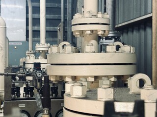 A close-up captures a detailed view of a large, beige-painted industrial flange connection. In the background, a complex assembly of pipes, valves, and gauges suggests a busy industrial or manufactur