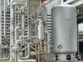 oil refinery plant. A close-up view of a large, silver industrial air receiver tank and a complex network of stainless steel pipes and valves at an industrial facility.