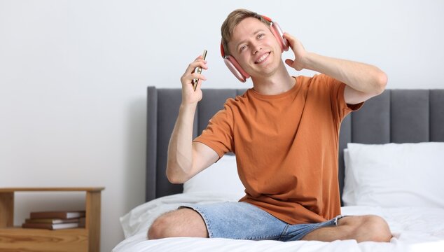Happy man with smartphone listening to music in headphones on bed at home