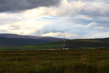 Remote industrial tower in northern Iceland surrounded by rolling hills and dramatic clouds, set in green and purple summer landscape, contrast of nature and human presence in wild Nordic terrain
