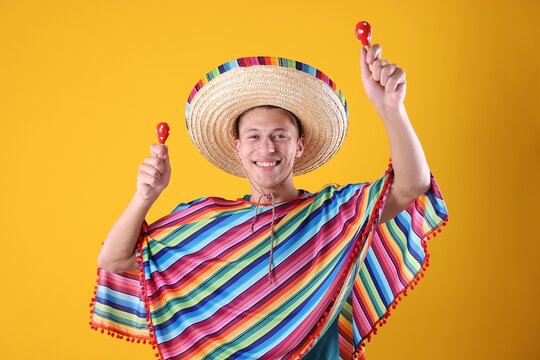 Happy man in sombrero hat and colorful poncho playing maracas on yellow background