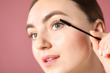 Young woman applying mascara onto her eyelashes on pink background, closeup