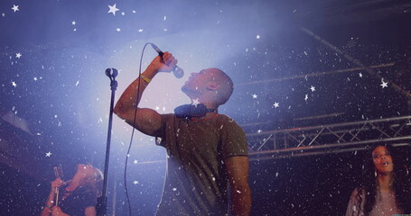 Singer wearing T-shirt singing on club stage under metal truss with headphones, mic and confetti