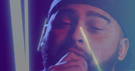 Singing bearded man wearing backward cap performing under neon tubes at concert, with microphone