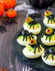 Obraz premium Halloween deviled eggs topped with black olive and green olive spiders, displayed on a round black slate with mini pumpkins, plastic spiders, and rustic wood background for a spooky party presentation