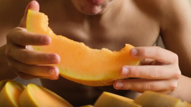 Front view portrait of a teenage boy biting a ripe juicy melon slice with both hands. Child loves fruit and eats with appetite. Hungry boy greedily enjoying melon indoors at the table.