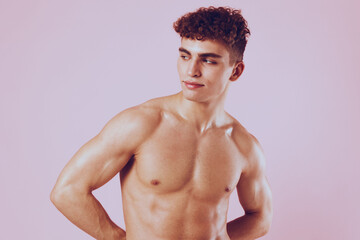 Fototapeta premium Confident young man with curly hair posing shirtless against a soft pink background, showcasing fitness and body positivity. Fashion and beauty concept.