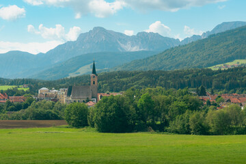 Charming alpine town photographed from a train, featuring a historic church with tall steeple, surrounded by green fields, red rooftops, and majestic mountain backdrop under a summer sky.