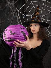 Beautiful witch girl in a witch's cap holds a lilac lantern in her hands against the background of a white web on Halloween