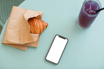 Overhead view of a mobile phone with empty screen mock-up, an iced blue matcha tonic drink and a croissant in a paper bag on a garden table in the summer