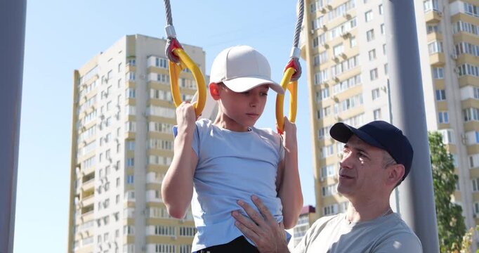 Father helps son exercise playground rings. Child trains with focus while dad supports with smile. Father helps kid exercise outdoors, showing care. Father helps child exercise with love. Happy family