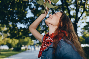 smiling woman enjoying nature outdoors in sunny park with green trees and casual denim jacket and red scarf accessory