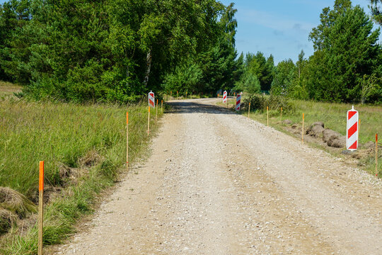 Gravel road under construction marked with stakes and warning signs in rural green landscape