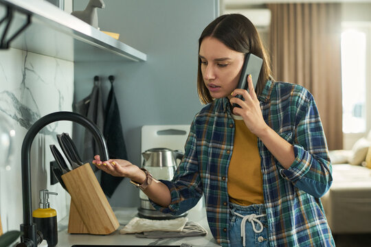 Young adult Caucasian woman standing in kitchen talking on smartphone while checking water from faucet, appearing concerned and examining hand under running tap - Powered by Adobe