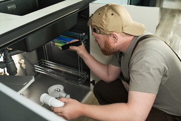 Caucasian middle aged man wearing cap kneeling under sink inspecting plumbing with flashlight holding pipe component focusing on repair task in modern kitchen environment