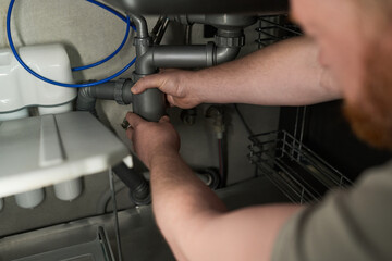 Obraz premium Caucasian middle aged man repairing kitchen sink plumbing under counter, using both hands to adjust pipe connections, focusing on maintenance and home improvement task