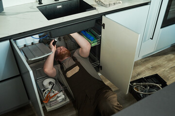 Caucasian young adult man lying under kitchen sink repairing plumbing using flashlight and tools, wearing work overalls, toolbox and pipes visible on floor beside open cabinet